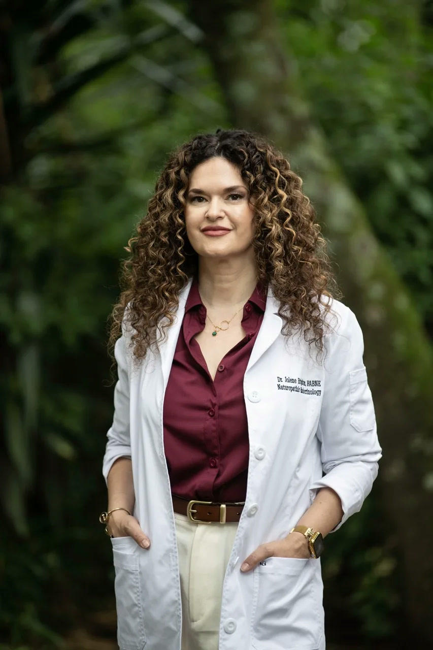Dr. Jolene Brighten in a white lab coat embroidered with her name and credentials, smiling in a botanical garden